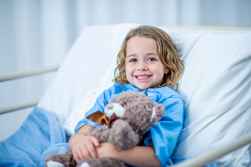A Caucasian girl is indoors in a hospital room. She is smiling at the camera while holding a teddy bear.
