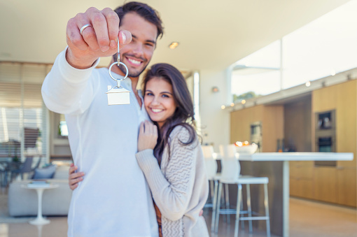 Couple holding a house key in their new home. They are standing in their new modern house. Both are happy and smiling. The house key has a house icon keyring