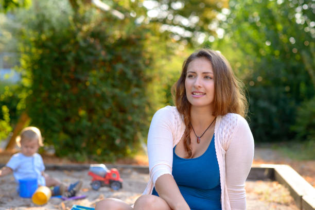 Thoughtful young mother playing outdoors with her baby son sitting looking to the side with a pensive expression against a background of greenery