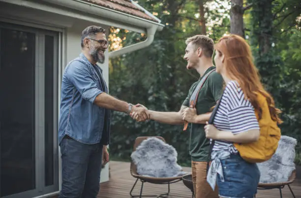 Young couple renting house. House owner shaking hands with couple who rent his house. Couple is on vacation and they are wearing backpacks.