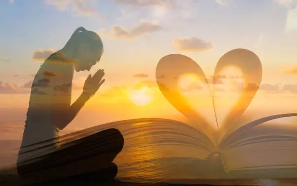 Young female praying next to bible with heart shape page against beautiful sunset.