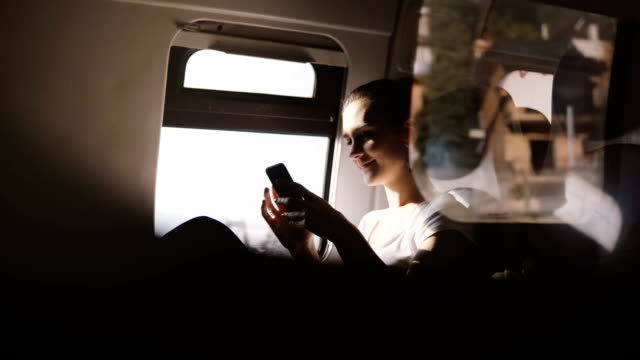 Amazing reflection shot of happy European girl smiling, using smartphone app sitting on modern moving train window seat