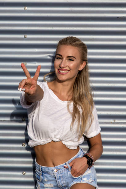 Potrait of a young blonde woman in front of a metallic wall A young blonde with a great, sporty figure stands on a metallic wall in the beautiful sunlight. She wears very short tight jeans, a white shirt and open hair. She enjoys the day and her life. She likes photography, analogue cameras, music and well-made sustainably produced coffee. She is in a good mood, in her mid-20s and very beautiful. sonnenbrille stock pictures, royalty-free photos & images