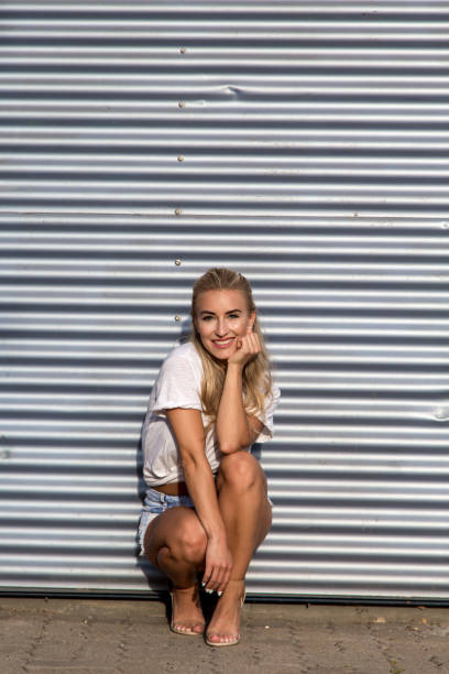 Potrait of a young blonde woman in front of a metallic wall A young blonde with a great, sporty figure stands on a metallic wall in the beautiful sunlight. She wears very short tight jeans, a white shirt and open hair. She enjoys the day and her life. She likes photography, analogue cameras, music and well-made sustainably produced coffee. She is in a good mood, in her mid-20s and very beautiful. sonnenbrille stock pictures, royalty-free photos & images