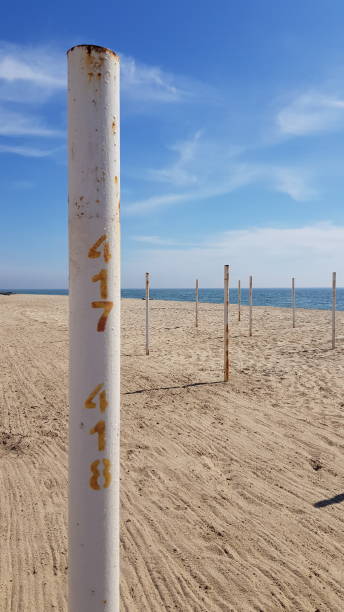 Desert beach landscape with white rusty pole and numbers on it. Minimalistic seascape. Beach with nobody. White sand and blue sea with white sky clouds above. Noon shadows on the beach. Tourism and vacation themed. Travel background.