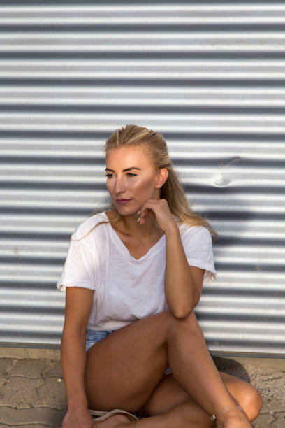 Potrait of a young blonde woman in front of a metallic wall A young blonde with a great, sporty figure stands on a metallic wall in the beautiful sunlight. She wears very short tight jeans, a white shirt and open hair. She enjoys the day and her life. She likes photography, analogue cameras, music and well-made sustainably produced coffee. She is in a good mood, in her mid-20s and very beautiful. sonnenbrille stock pictures, royalty-free photos & images