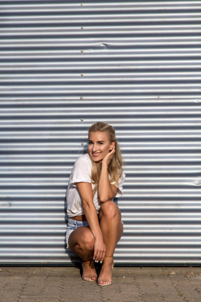 Potrait of a young blonde woman in front of a metallic wall A young blonde with a great, sporty figure stands on a metallic wall in the beautiful sunlight. She wears very short tight jeans, a white shirt and open hair. She enjoys the day and her life. She likes photography, analogue cameras, music and well-made sustainably produced coffee. She is in a good mood, in her mid-20s and very beautiful. sonnenbrille stock pictures, royalty-free photos & images