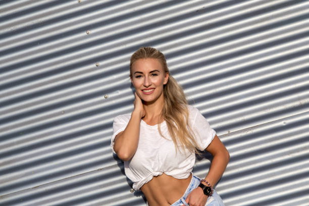 Potrait of a young blonde woman in front of a metallic wall A young blonde with a great, sporty figure stands on a metallic wall in the beautiful sunlight. She wears very short tight jeans, a white shirt and open hair. She enjoys the day and her life. She likes photography, analogue cameras, music and well-made sustainably produced coffee. She is in a good mood, in her mid-20s and very beautiful. sonnenbrille stock pictures, royalty-free photos & images