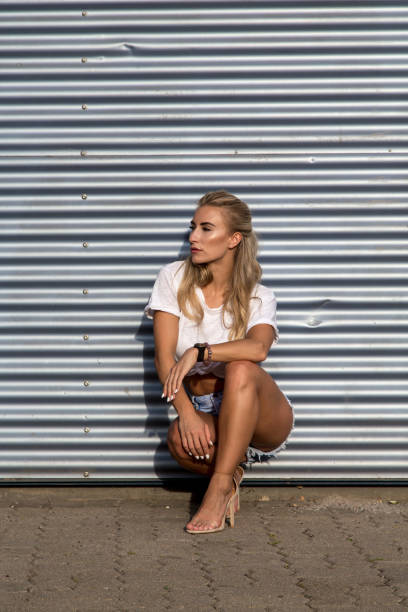 Potrait of a young blonde woman in front of a metallic wall A young blonde with a great, sporty figure stands on a metallic wall in the beautiful sunlight. She wears very short tight jeans, a white shirt and open hair. She enjoys the day and her life. She likes photography, analogue cameras, music and well-made sustainably produced coffee. She is in a good mood, in her mid-20s and very beautiful. sonnenbrille stock pictures, royalty-free photos & images