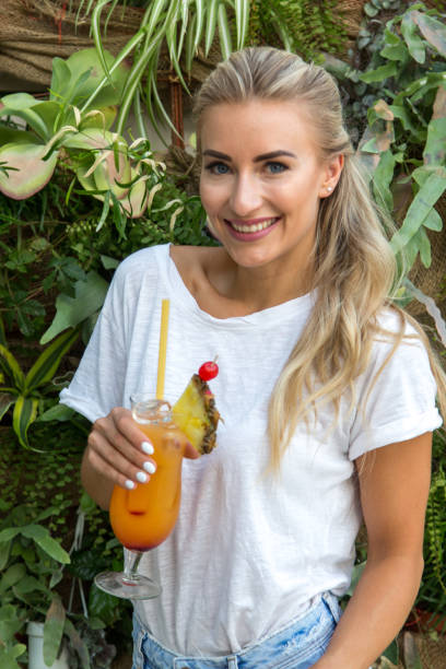 Young woman drinks a cocktail A young blonde with a great, sporty figure stands on a metallic wall in the beautiful sunlight. She wears very short tight jeans, a white shirt and open hair. She enjoys the day and her life. She likes photography, analogue cameras, music and well-made sustainably produced coffee. She is in a good mood, in her mid-20s and very beautiful. sonnenbrille stock pictures, royalty-free photos & images