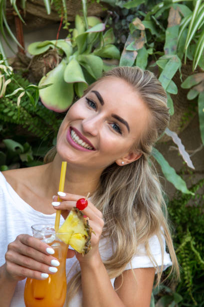 Young woman drinks a cocktail A young blonde with a great, sporty figure stands on a metallic wall in the beautiful sunlight. She wears very short tight jeans, a white shirt and open hair. She enjoys the day and her life. She likes photography, analogue cameras, music and well-made sustainably produced coffee. She is in a good mood, in her mid-20s and very beautiful. sonnenbrille stock pictures, royalty-free photos & images