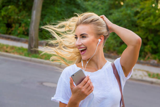 Potrait of a young blonde woman listening to the music and dancing A young blonde with a great, sporty figure stands on a metallic wall in the beautiful sunlight. She wears very short tight jeans, a white shirt and open hair. She enjoys the day and her life. She likes photography, analogue cameras, music and well-made sustainably produced coffee. She is in a good mood, in her mid-20s and very beautiful. sonnenbrille stock pictures, royalty-free photos & images