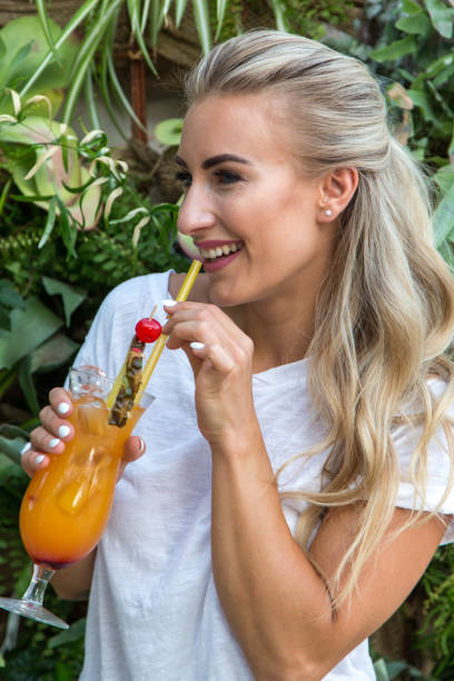 Young woman drinks a cocktail A young blonde with a great, sporty figure stands on a metallic wall in the beautiful sunlight. She wears very short tight jeans, a white shirt and open hair. She enjoys the day and her life. She likes photography, analogue cameras, music and well-made sustainably produced coffee. She is in a good mood, in her mid-20s and very beautiful. sonnenbrille stock pictures, royalty-free photos & images