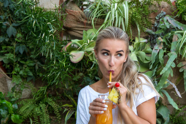 Young woman drinks a cocktail A young blonde with a great, sporty figure stands on a metallic wall in the beautiful sunlight. She wears very short tight jeans, a white shirt and open hair. She enjoys the day and her life. She likes photography, analogue cameras, music and well-made sustainably produced coffee. She is in a good mood, in her mid-20s and very beautiful. sonnenbrille stock pictures, royalty-free photos & images