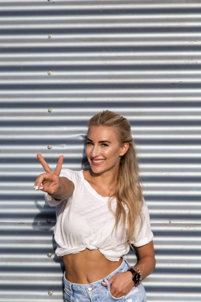 Potrait of a young blonde woman in front of a metallic wall A young blonde with a great, sporty figure stands on a metallic wall in the beautiful sunlight. She wears very short tight jeans, a white shirt and open hair. She enjoys the day and her life. She likes photography, analogue cameras, music and well-made sustainably produced coffee. She is in a good mood, in her mid-20s and very beautiful. sonnenbrille stock pictures, royalty-free photos & images