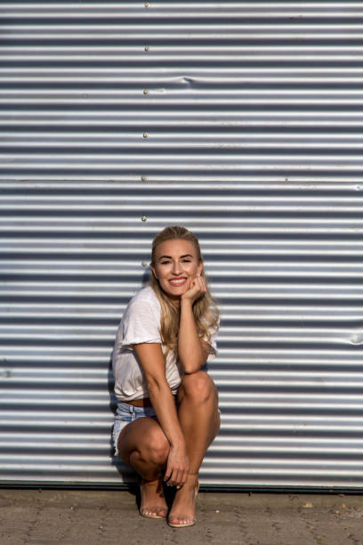 Potrait of a young blonde woman in front of a metallic wall A young blonde with a great, sporty figure stands on a metallic wall in the beautiful sunlight. She wears very short tight jeans, a white shirt and open hair. She enjoys the day and her life. She likes photography, analogue cameras, music and well-made sustainably produced coffee. She is in a good mood, in her mid-20s and very beautiful. sonnenbrille stock pictures, royalty-free photos & images