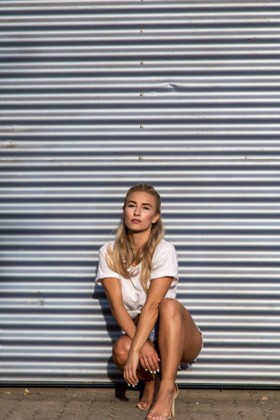 Potrait of a young blonde woman in front of a metallic wall A young blonde with a great, sporty figure stands on a metallic wall in the beautiful sunlight. She wears very short tight jeans, a white shirt and open hair. She enjoys the day and her life. She likes photography, analogue cameras, music and well-made sustainably produced coffee. She is in a good mood, in her mid-20s and very beautiful. sonnenbrille stock pictures, royalty-free photos & images