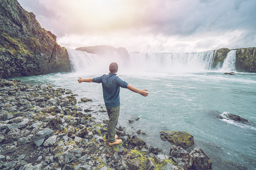 Young man arms outstretched in front of the magnificent waterfall in Iceland, Godafoss falls. People travel exploration concept