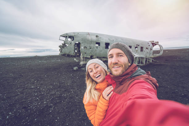 jovem casal em pé por acidente de avião na praia de areia preta, tomando um lugar famoso retrato de selfie para visitar na islândia e posar com o naufrágio - acidente evento relacionado com o transporte - fotografias e filmes do acervo