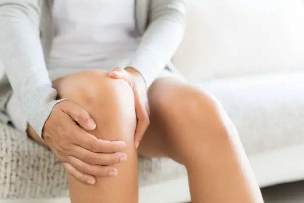 Photo of Closeup young woman sitting on sofa and feeling knee pain and she massage her knee at home. Healthcare and medical concept.