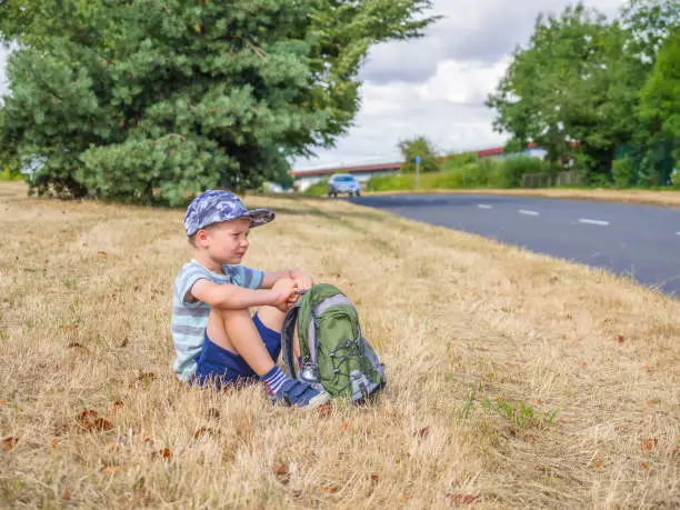 Photo of Day view little child boy traveler with backpack sitting next to UK road