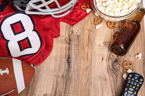 Football season is here.  Concept of sports fan watching the game on TV at home, at tailgate party, or sports bar with snacks and drinks.  TV remote, popcorn, football, jersey, helmet and beer bottle.