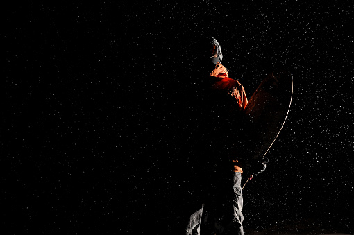 Bottom view of darkened figure of a male snowboarder standing on the snow at the dark snowy night