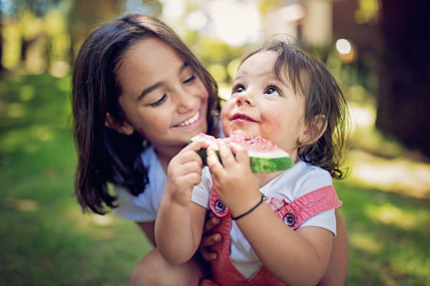 девушка обнимает свою младшую сестру, которая ест арбуз - healthy lifestyle people eating sister стоковые фото и изображения