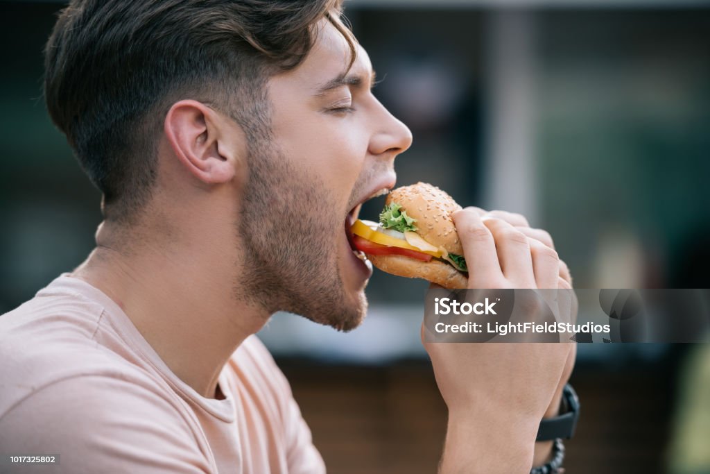 Vista lateral de homem comendo hambúrguer saboroso com os olhos fechados - Foto de stock de Comer royalty-free