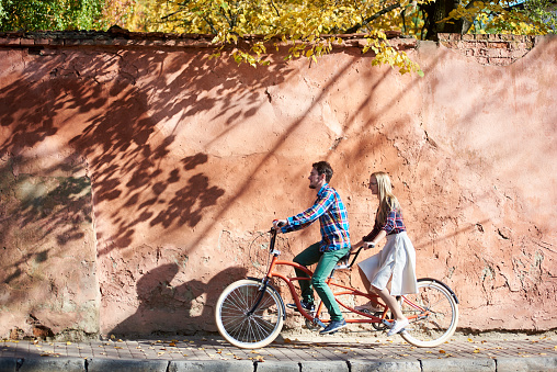 Young attractive happy tourist couple in casual clothing, bearded man and blond woman in glasses cycling together on double tandem bike on background of high red old crackled plastered wall.