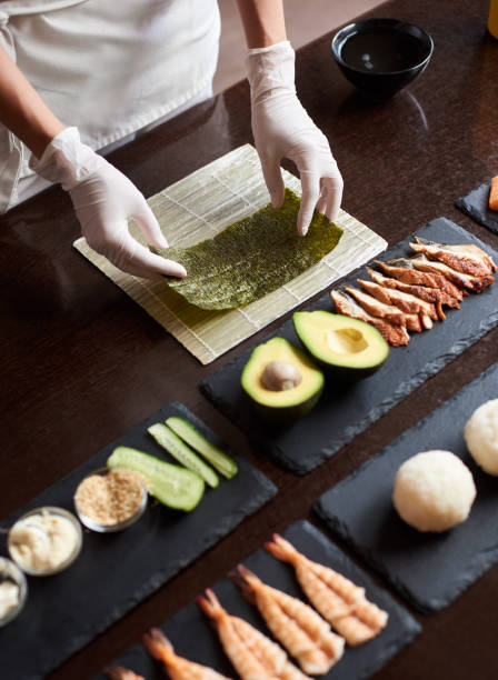 Process of preparing rolling sushi Closeup view of process of preparing rolling sushi. Chef's hands are holding sheet of nori. Ingredients: shrimp, cucumber, salmon, rice, avocado on black stone plates nori edible seaweed sheet stock pictures, royalty-free photos & images