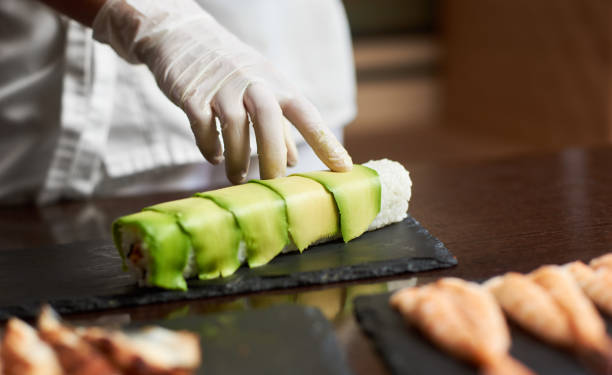Process of preparing rolling sushi Close-up view of process of preparing rolling sushi at. Hand in glove decorates roll with sliced avocado food state preparation shrimp prepared shrimp stock pictures, royalty-free photos & images