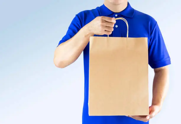 Photo of Delivery man in blue uniform and holding paper bag with delivering package on gray background. Concept fast food delivery service or order online shopping and express delivery.
