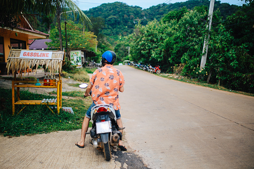 Young man riding a motorcycle on the island of Koh Lanta, Thailand in the Krabi province. He is wearing a helmet and sunglasses, enjoying his day, traveling solo through the beautiful nature of the islands in Thailand. He just made a break, stopping to buy some more gasoline for the trip.