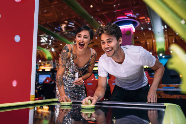 Happy couple playing coin operated air hockey game at a gaming parlour Couple playing air hockey game holding strikers at a gaming parlour. Excited man and woman having fun playing games at a gaming arcade. arcade photos stock pictures, royalty-free photos & images