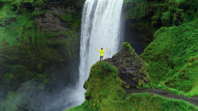 Aerial drone shot of man with arms raised on top of cliff standing in front of waterfall celebrating
