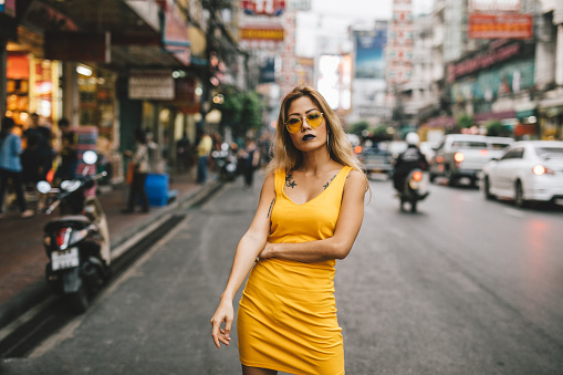 Portrait of fashionable Asian woman in the streets of bangkok. She's wearing a yellow dress and is looking at the camera.