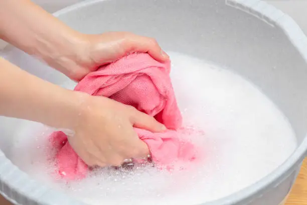 Photo of Woman hand washing knitted woolen laundry in grey plastic basin.Lot of soap white detergent. Dry skin and irritation