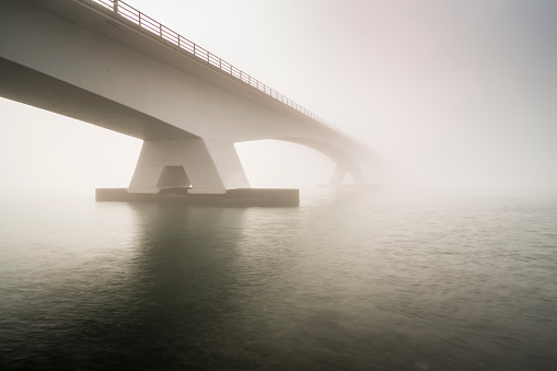 Zeeland Bridge (Zeelandbrug) covered in fog during sunrise in winter. Low visibilty making the sun shining through the fog  The Zeeland bridge is the longest bridge in the Netherlands, loved by tourists who visit Zeeland.