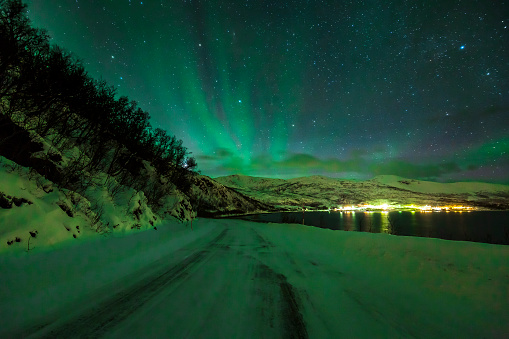 Northern lights Aurora Borealis in the night. A arctic, snowy winter landscape with a road and fjord on the foreground. The sky is clear, stars are visible