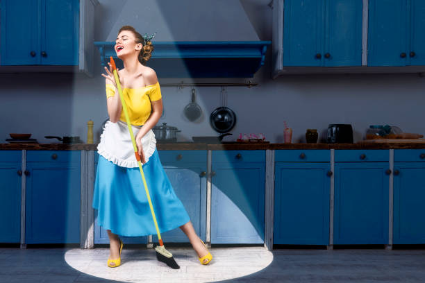 Retro pin up housewife on kitchen Retro pin up girl woman female housewife wearing yellow top, blue skirt and white apron holding mop singing and cleaning floor in stage light kitchen with blue cabinets and utensils. Housework concept retro 1950s kitchen stock pictures, royalty-free photos & images