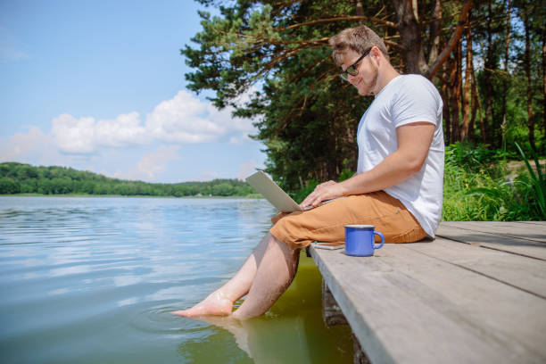 istock man working on laptop while sitting on wooden dock. legs in river water 1018044038
