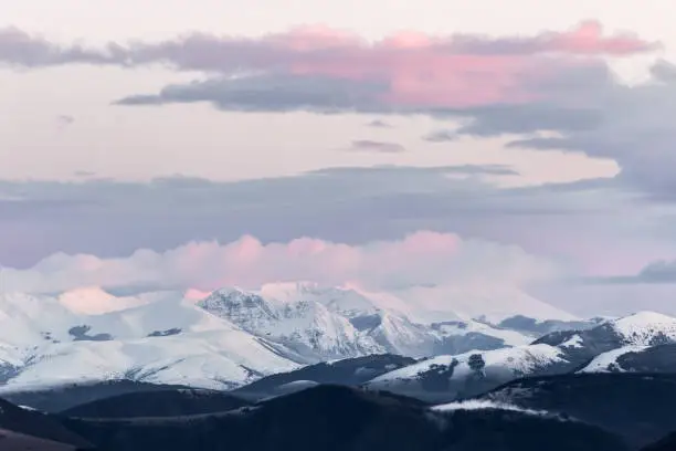 Photo of Mountain range covered by snow at sunset, with warm, purple shades reflections