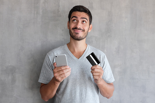 Hombre guapo joven pensando en compras en línea a través de internet, con camiseta gris, de pie contra la pared con textura, con tarjeta de crédito y teléfono celular photo
