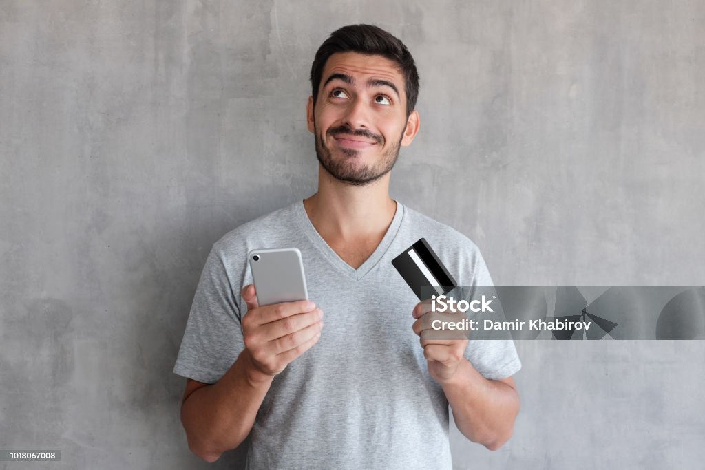 クレジット カードや携帯電話を保持している織り目加工の壁に立っているグレーの t シャツを着て、インターネット経由でオンライン ショッピングを考えて若いハンサムな男 - 男性のロイヤリティフリーストックフォト