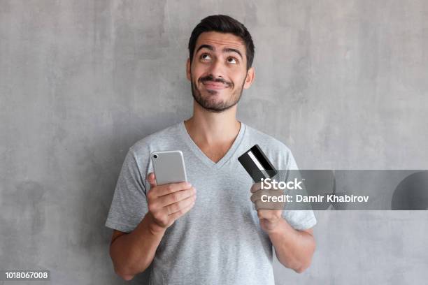 잘생긴 젊은이 인터넷 질감된 벽에 서 있는 회색 티셔츠를 입고 신용 카드 및 휴대폰을 통해 온라인 쇼핑에 대 한 생각 남자에 대한 스톡 사진 및 기타 이미지
