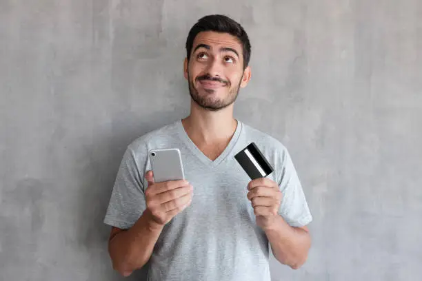 Photo of Young handsome man thinking about online shopping via internet, wearing gray t shirt, standing against textured wall, holding  credit card and cell phone