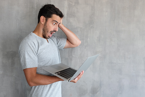 Handsome surprised man in gray t shirt looking at the laptop screen, astonished with big sale prices, holding hand on head, standing against textured wall with copy space