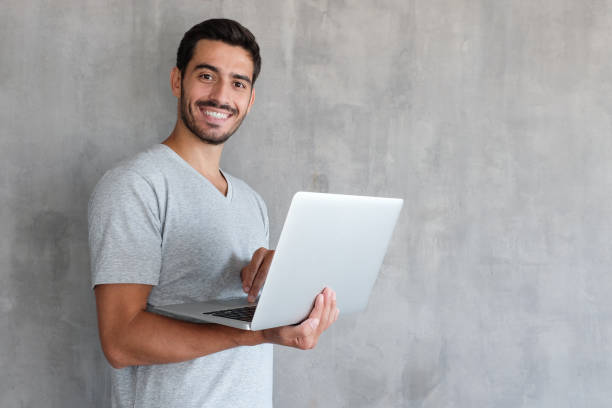 indoor-porträt von junger mann in t-shirt gegen strukturierte wand mit textfreiraum für anzeigen, laptop halten und blick in die kamera mit glücklichen lächeln - männliche figur stock-fotos und bilder