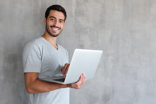 Indoor portrait of young man in t shirt standing against textured wall with copy space for ads, holding laptop and looking at camera with happy smile